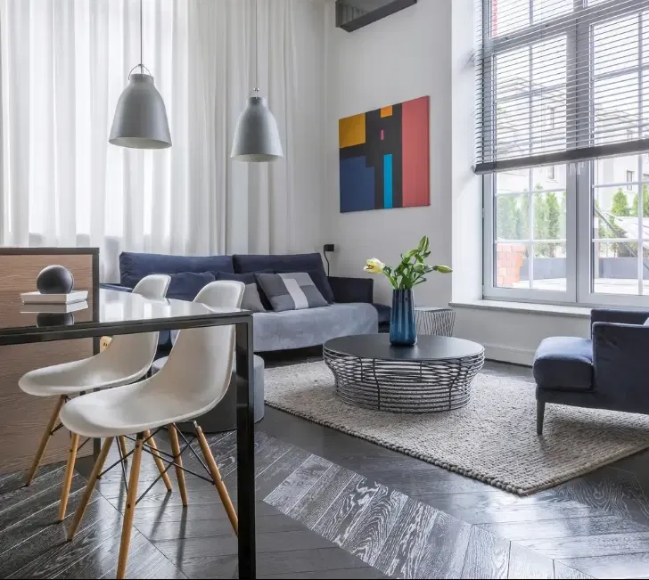 Salon lumineux avec canapé, fauteuil et grande fenêtre avec stores.